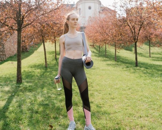 Athletic woman holding water bottle and mat, posing outdoors in sunny park.