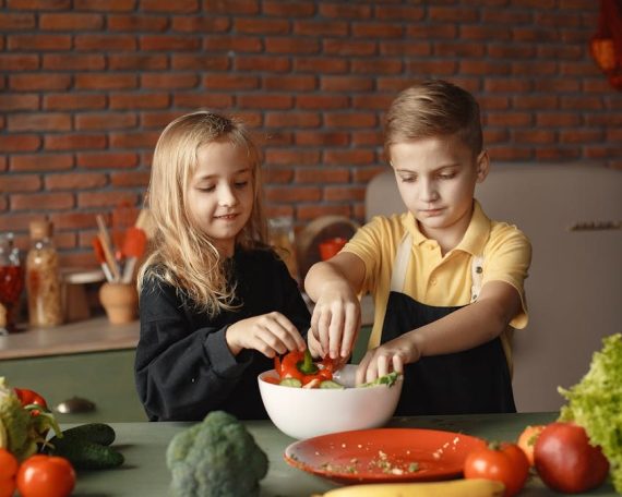 Cheerful girl helping little boy in apron preparing salad while standing in kitchen near table with mix of fresh vegetables behind brick wall